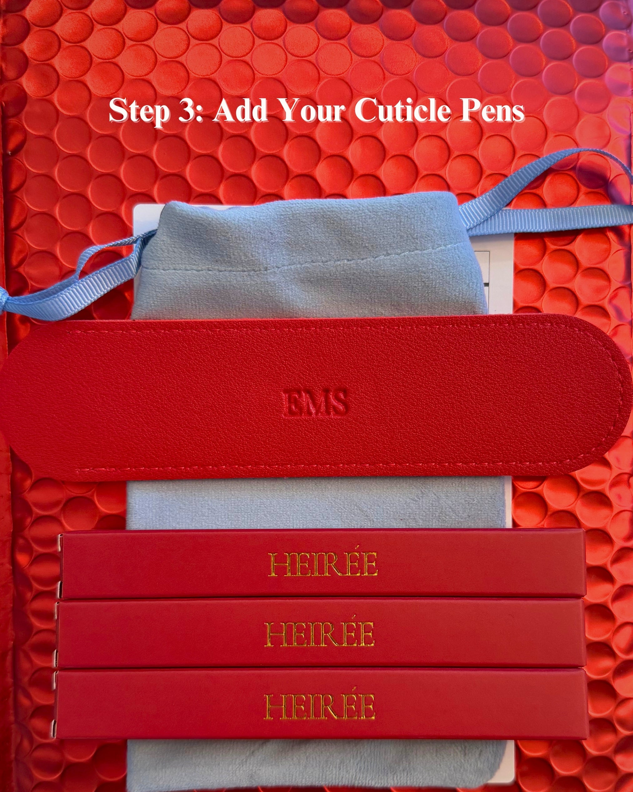 A close-up of a red leather cuticle tool set with three red bars engraved with the name HEIRÉE, a light blue velvet pouch, and a red strap, arranged on a textured red background with white text reading 'Step 3: Add Your Cuticle Pens'.
