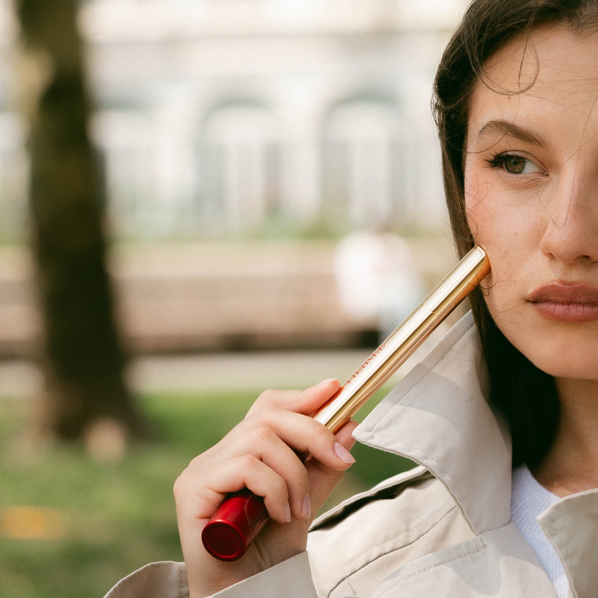 A close-up lifestyle shot of a woman holding a gold and red cosmetic tube against her cheek, wearing a beige trench coat outdoors with a blurred park background.