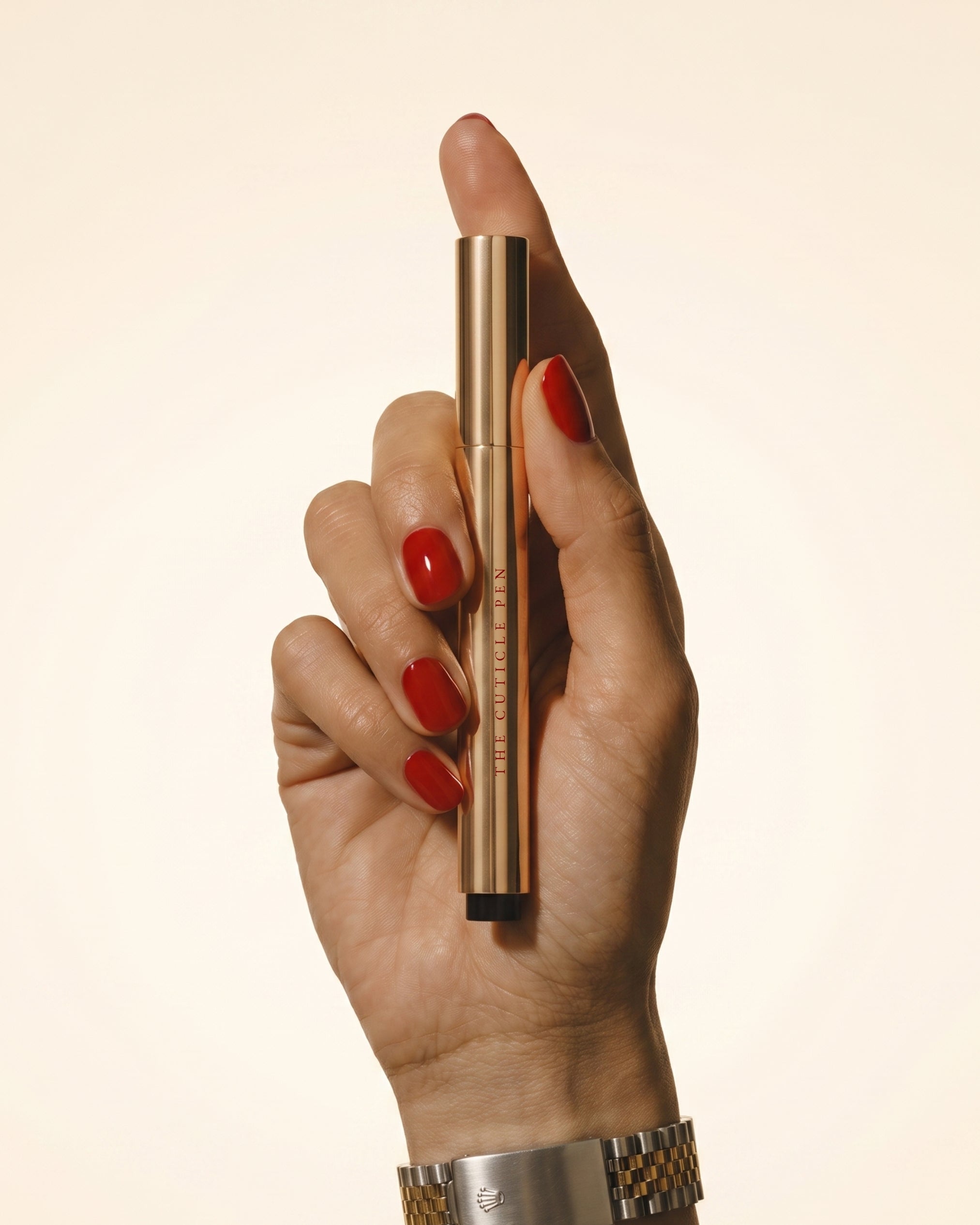 A hand with red nail polish holds a slim gold cosmetic tube vertically against a plain warm beige background. The tube has vertical text that appears to read 'THE SCIENCE OF MAKEUP' and is shown close up with a metal watch band visible at the wrist.
