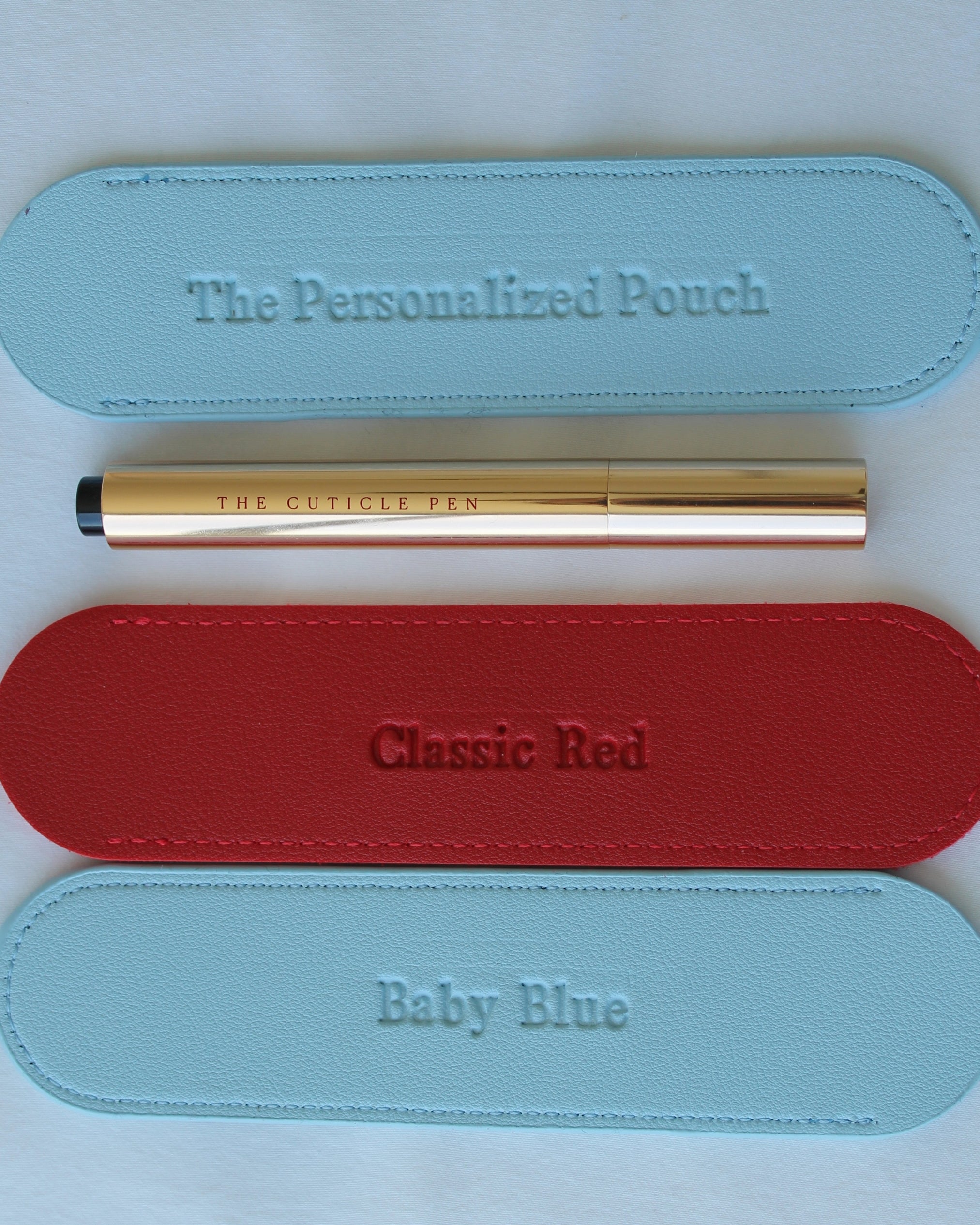 Three oval leather pouches in light blue, red, and light blue are arranged vertically on a white background, each with matching embossed text reading 'The Personalized Pouch,' 'Classic Red,' and 'Baby Blue.' A gold pen labeled 'THE CUTICLE PEN' lies horizontally across the center between the pouches.