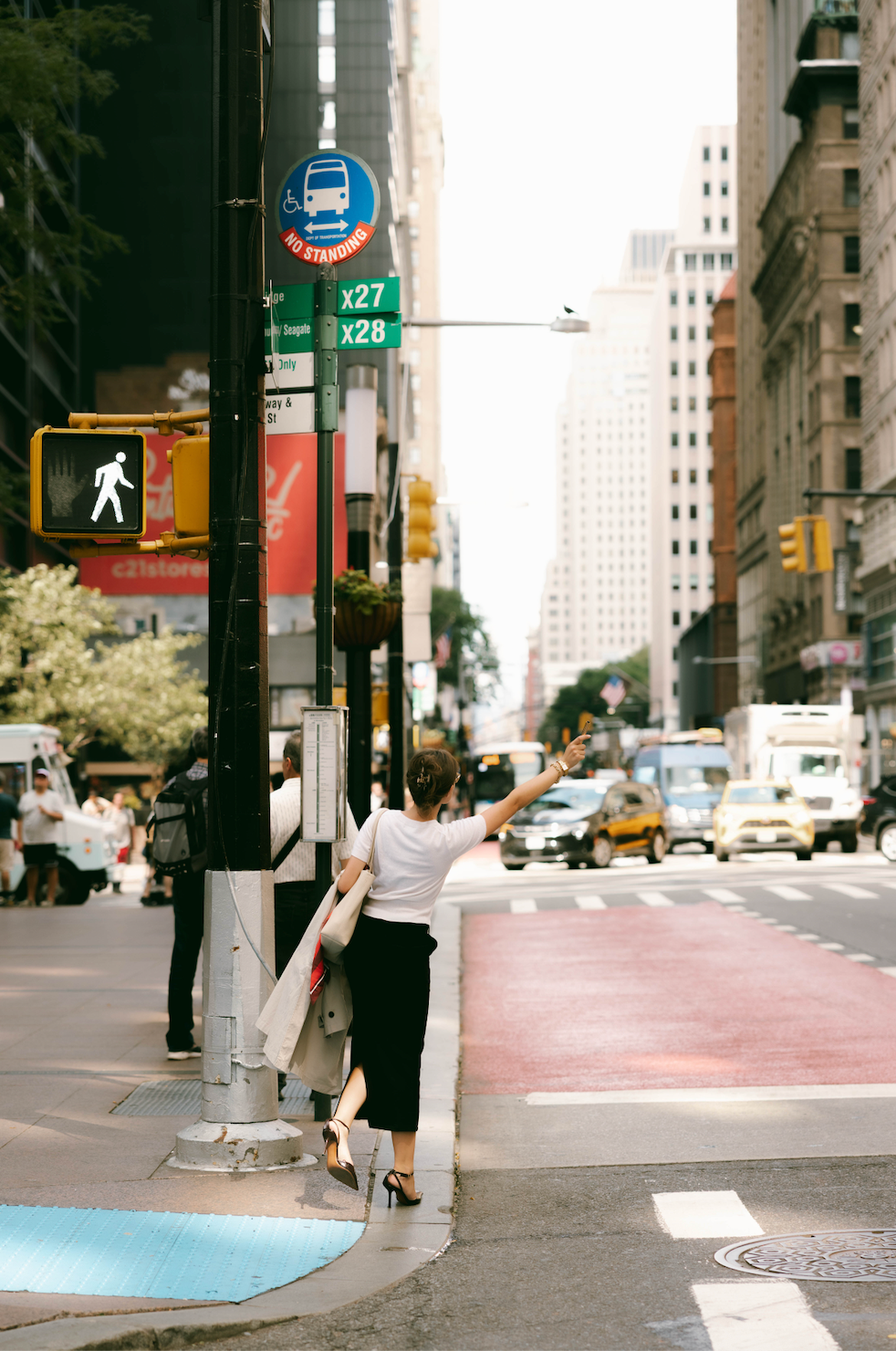 A woman in a white t-shirt and black skirt stands at a city crosswalk, reaching out to signal a bus. The New York City street scene features yellow cabs, tall buildings, traffic lights, and a red crosswalk under a bright sky.